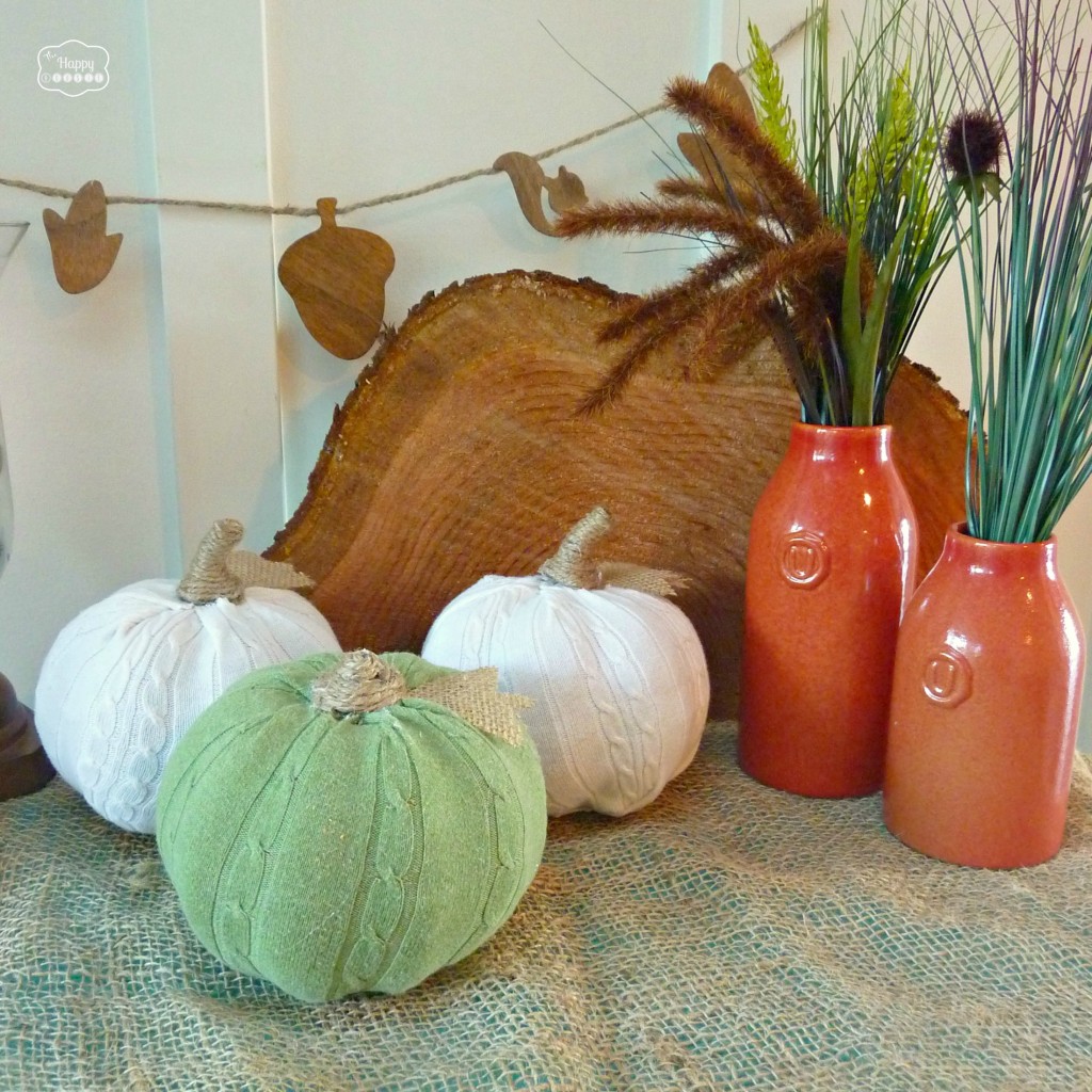Easy DIY Sweater Pumpkins with twine stems and burlap leaves on the counter beside some orange pottery.